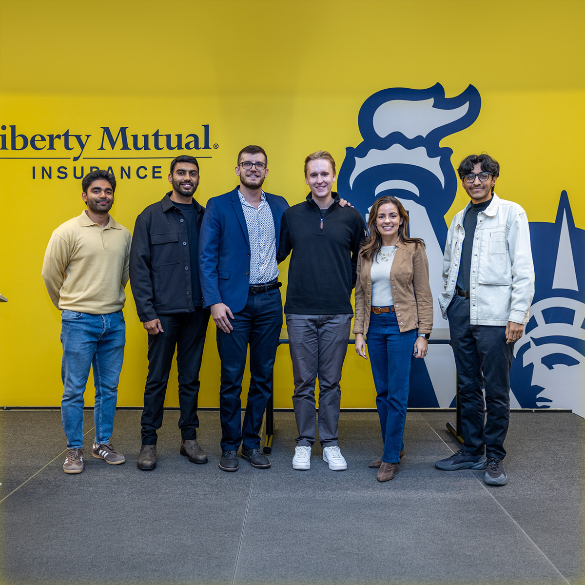 Ignite Hackathon participants stand with Monica Caldas in front of a large yellow wall with the Liberty Mutual medallion on it. 
